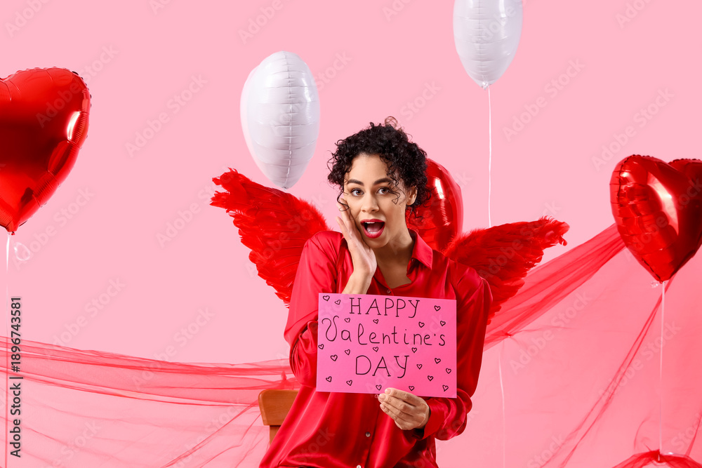 Fototapeta premium Surprised young African-American woman with Cupid's wings and card for Valentine's Day on pink background