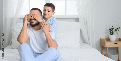 Happy son in pajamas closing his father's eyes on bed at home