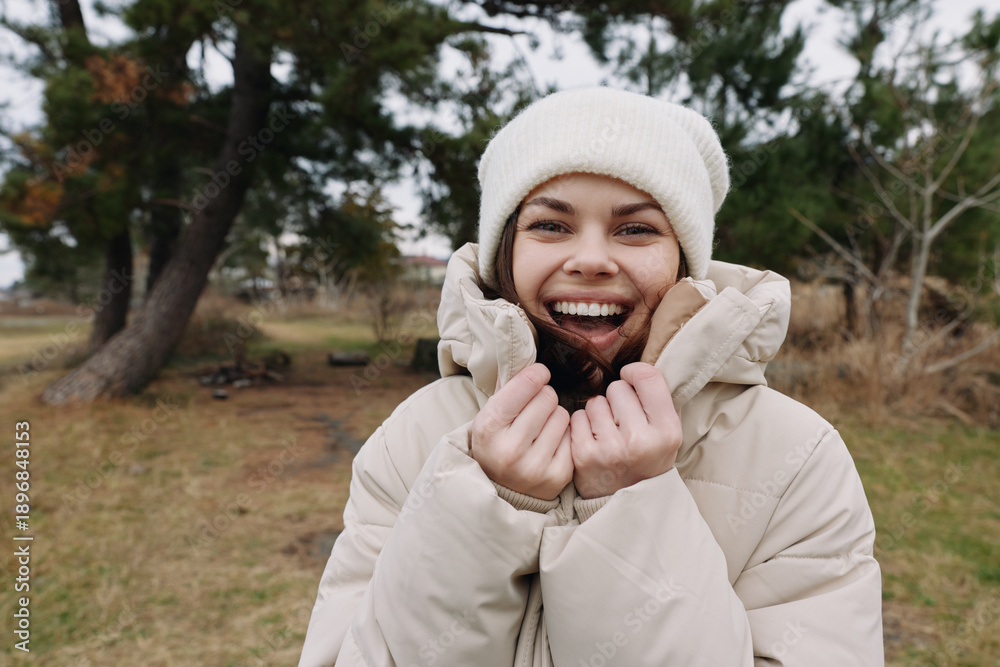 Obraz premium Woman outdoors wearing a warm winter coat and knit hat, smiling brightly in a park setting with trees and grass, casual cold weather moment.