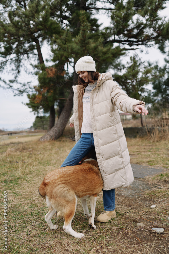 Obraz premium Woman in a long beige winter coat and beanie walks with a large dog in a park, grass and pine trees in the background, candid outdoor moment of companionship and exploration