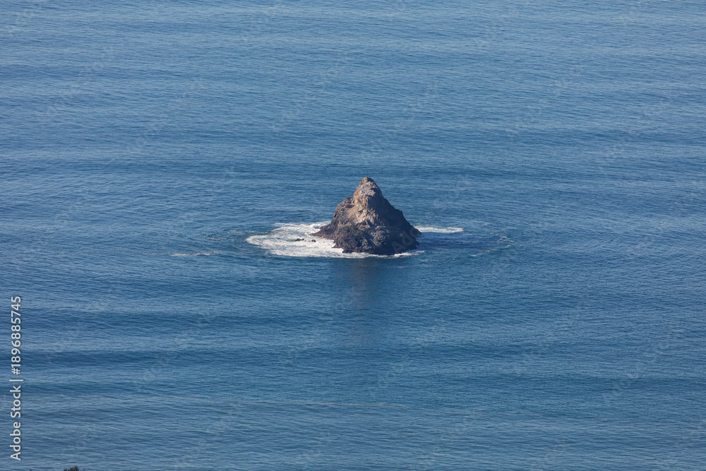 Fototapeta premium Small rocky sea stack in the Pacific Ocean off the Oregon Coast near Cannon Beach, USA surrounded by blue water and gentle waves in a calm natural coastal scene.