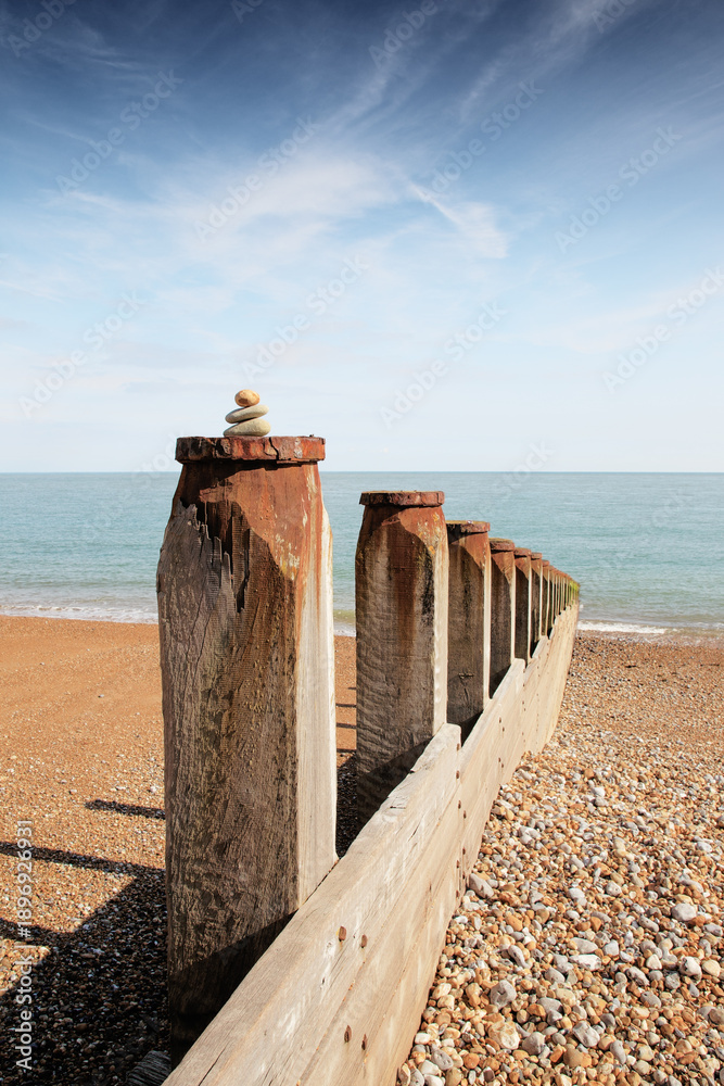 Fototapeta premium Coastal Wooden Breakwater With Stacked Stones on Pebble Beach Under Clear Blue Sky