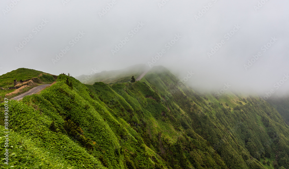 Naklejka premium Sete Cidades volcanic landscape in San Miguel, Azores. Lush green hillsides covered in fog and mist. Scenic view of the natural environment. Remote outdoor setting with winding path.