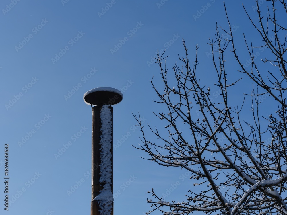 Naklejka premium Snow covered old metal chimney and tree branches against a blue sky.