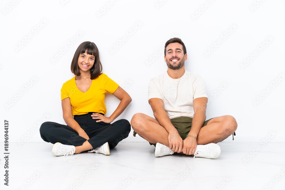 Fototapeta premium Young couple sitting on the floor isolated on white background posing with arms at hip and smiling