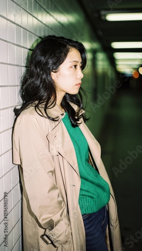 Young Asian woman in a trench coat leaning on a wall in a dark tunnel