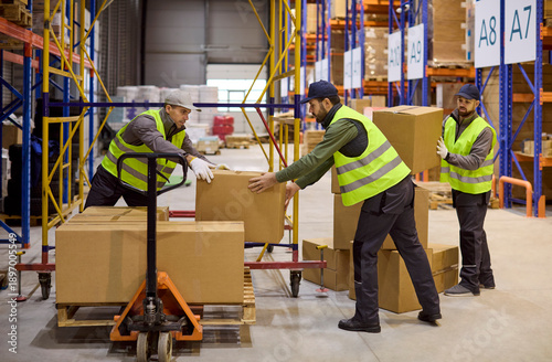 Workers in warehouse loading boxes on pallet. Loaders prepare shipment with pallet jack and stack parcels. Safety vests and logistics ensure fast order flow. Concept of teamwork and logistics.