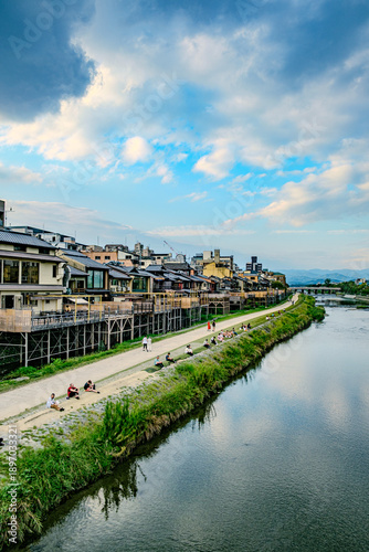 People relaxing on the bank of Kamo River in Kyoto with traditional buildings and clouds.Kyoto, Japan, Kamo River, Kamogawa, Riverbank, Relaxing, People, Travel, Summer, Evening, Traditional architect
