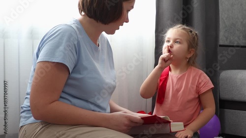 Mid shot of young Caucasian woman speech therapist reads book with cute girl. Mother and daughter learning together. Preschool education for young children.