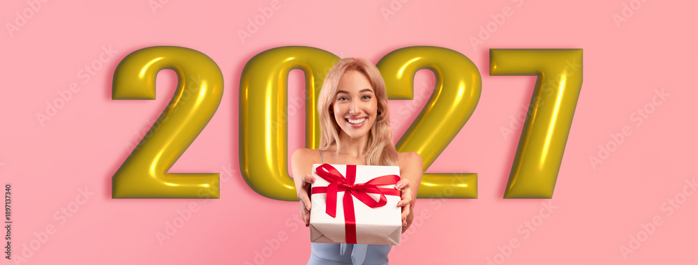 Fototapeta premium A woman stands smiling and holding a wrapped gift. Behind her are large numbers showing the year 2027. The background is pink. She appears happy and excited about the gift.