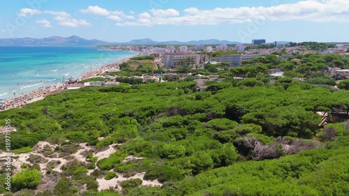 Aerial view of Playa de Muro beach in Mallorca, Spain, showcasing vibrant turquoise waters, sandy shoreline, lush greenery, and coastal buildings under a bright blue sky