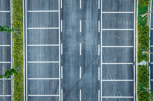 Aerial top view of an empty asphalt parking lot with white painted line and green hedge in an urban area under bright day.