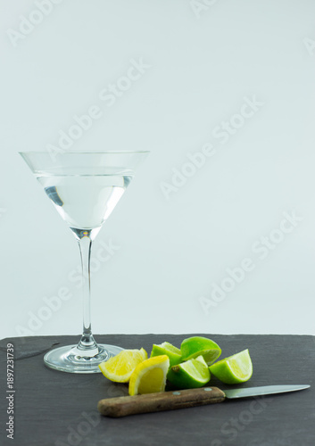A martini in a traditional martini glass set against a white background with wedges of lemons and limes, a small knife on a grey slate chopping board against a white background.