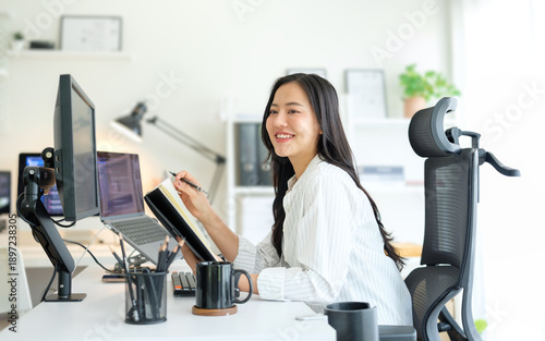 Slika na platnu Smiling female programmer working at a modern desk with dual monitors and laptop, writing notes while coding in a bright home office