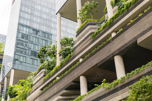 A building with multiple levels shows green terraces filled with various plants. This structure stands in a busy urban area surrounded by glass buildings