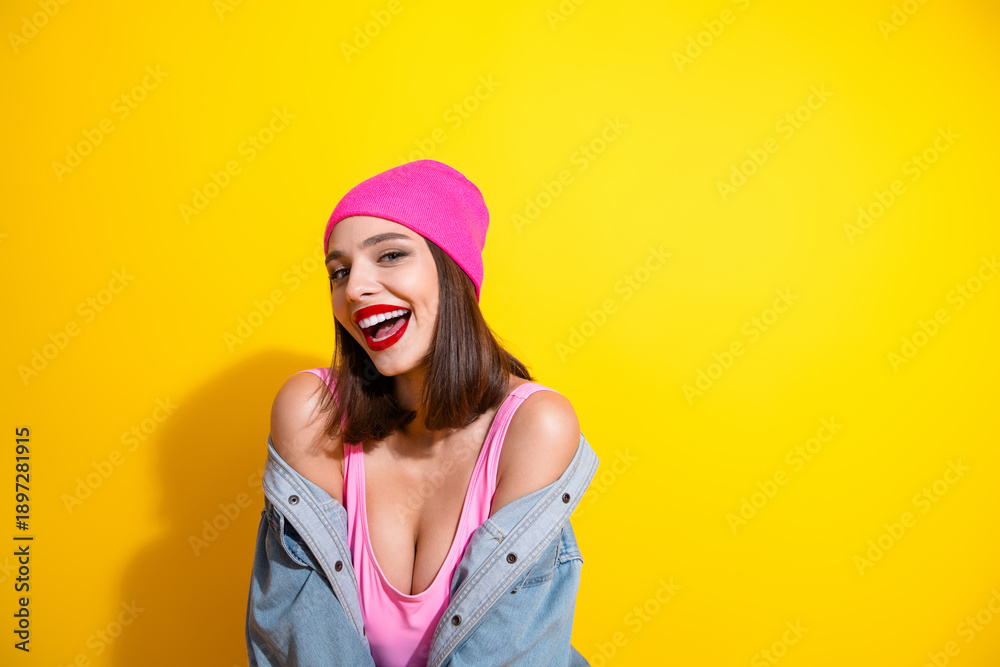 Naklejka premium Young woman in pink beanie laughs in a bright studio against yellow background fashion shoot