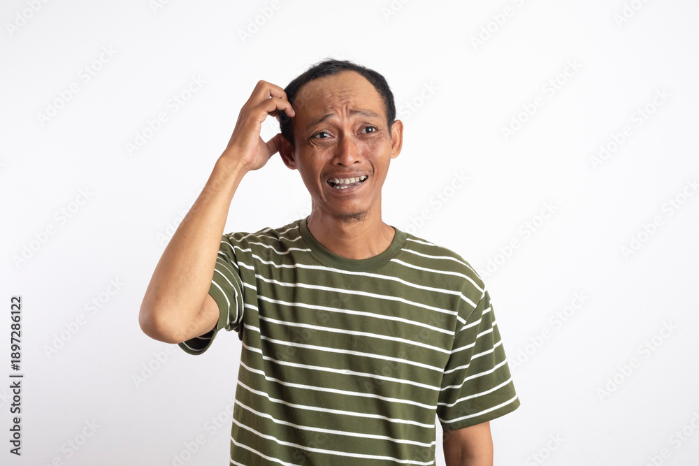 Fototapeta premium Confused Asian man scratching his head while looking up with an awkward smile. Casual studio portrait on white background representing doubt, uncertainty, thinking process, hesitation, 