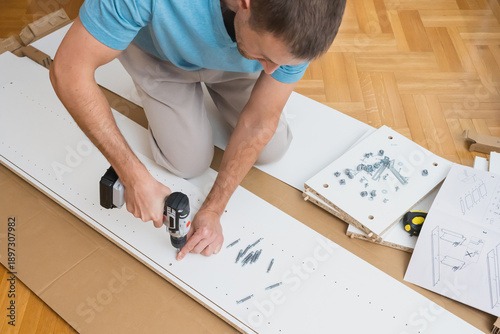 Male hands use a screwdriver to fasten hardware into a white furniture panel with assembly parts and instructions