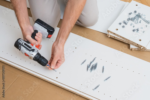 Male hands use a screwdriver to fasten hardware into a white furniture panel with assembly parts and instructions