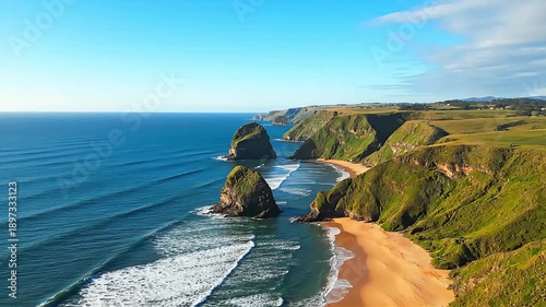 Dramatic ocean waves crash against rugged green cliffs and sandy beach under a clear blue sky.