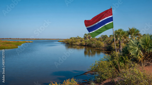 A Gambian flag is flying by a river with clear water. Lush green trees and tropical plants are visible along the bank. The scene is bright and captures the beauty of nature in this location.
