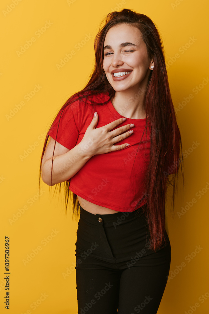 Obraz premium Young woman with long hair smiling and winking with hand on chest wearing red shirt and black pants standing against yellow background. Positive emotion and casual style portrait.