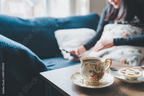 Relaxing morning scene or afternoon tea with teacup,sweet pie on coffee table,Blurry woman use smartphone resting on sofa in background,Concept slow life,mental health,quiet moment of wellness at home