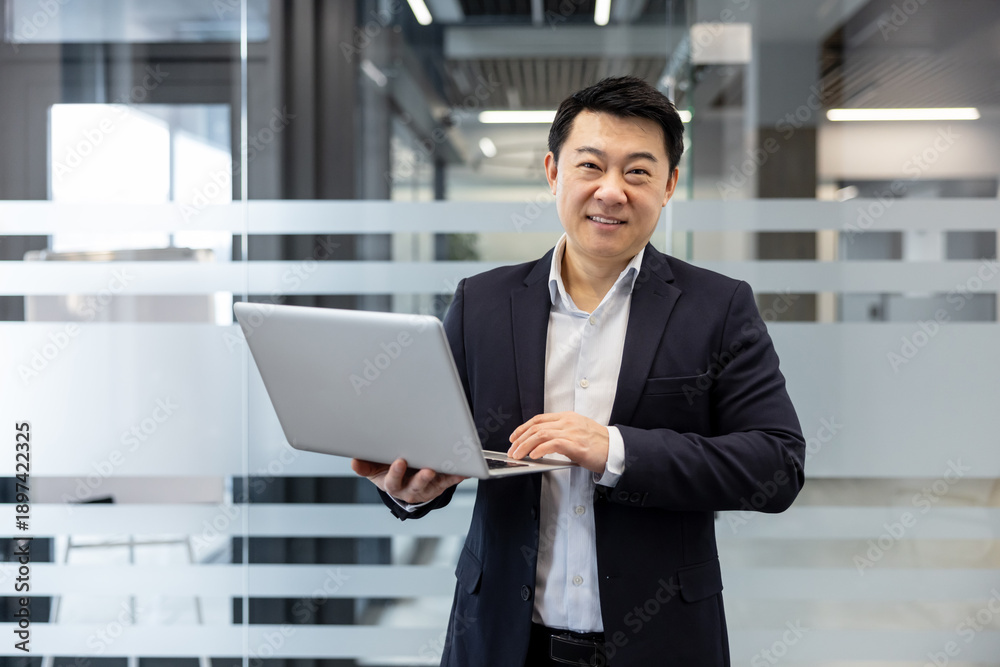 Fototapeta premium Asian businessman in a suit smiling confidently while typing on a silver laptop in a bright, modern office hallway with frosted glass partitions and clean architectural lines