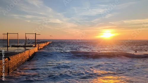 Sea at sunrise with golden sun shining on waves rolling on shore with pier