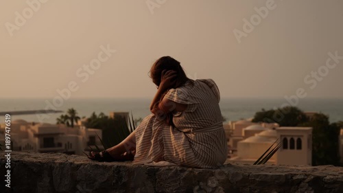A woman in a striped dress sits on a stone wall overlooking the sea and traditional buildings. The warm sunset light creates a serene and reflective atmosphere. Woman relaxes on a stone ledge