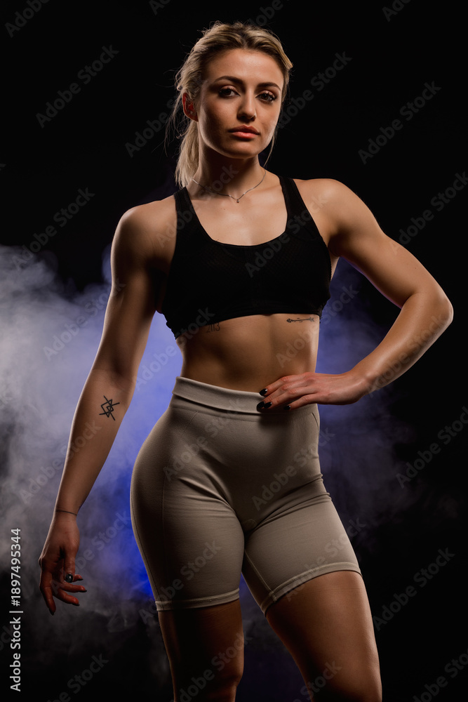 Fototapeta premium Confident athletic woman in black sports bra and shorts posing against dark smoky background exuding strength and empowerment