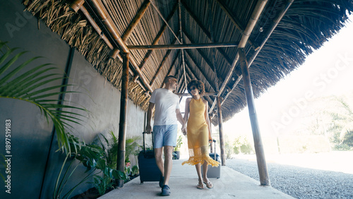 Multiracial couple holding hands, smiling while walking at a tropical resort, arriving for vacation with their bags