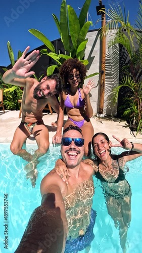 Four happy friends having fun together in a swimming pool on a sunny summer day. A young man records a selfie video smiling at the camera. Vertical