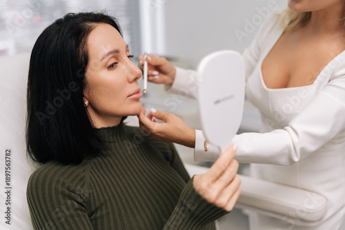 Foto Beautiful client consulting with cosmetologist about potential facial aesthetic treatments, looking in mirror while professional explains options and marks areas for improvement in beauty clinic