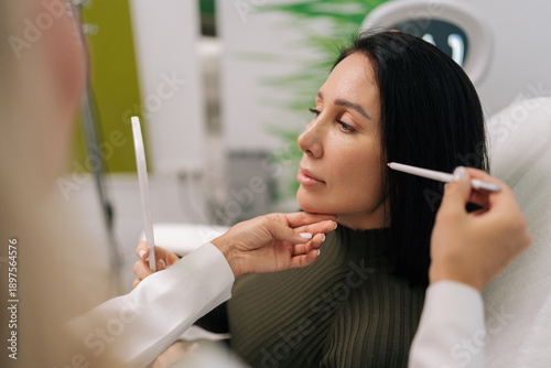Konstfotografi Side view of woman looking in mirror while cosmetologist marking face for aesthetic procedure, preparing for beauty treatment in cosmetology clinic