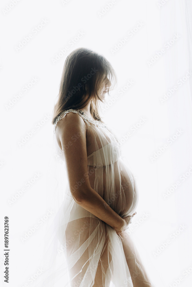 Obraz premium Beautiful pregnant woman holding baby bump and posing in light. Happy Mothers day! Healthy pregnancy and fertility concept. Pregnant woman in veil hugging belly, tender motherhood