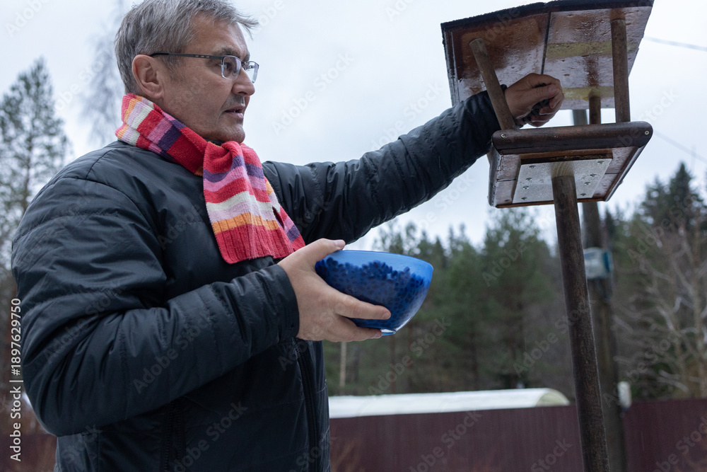 Φωτογραφία Man refilling bird feeder with seeds in snowy garden wearing jacket and scarf among trees during cold season