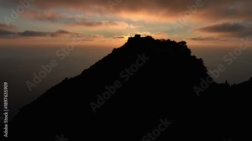 Aerial view of the sun rising from behind the mountain peak silhouette of Angelokastro castle, casting golden light over the sea, Corfu, Greece