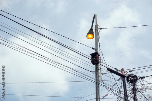 Obraz na płótnie This electric pole lamp is lit during the day under a cloudy sky, surrounded by many messy and tangled electric cables