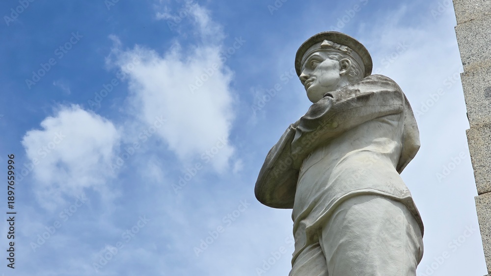Obraz premium Maryborough, Australia - 4 January 2026. A carved stone soldier at the Maryborough War Memorial faces outward with arms crossed, set against a blue sky with scattered clouds.