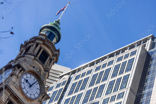 Photography Historic sandstone clock tower with Australian flag beside modern glass office building under clear blue sky