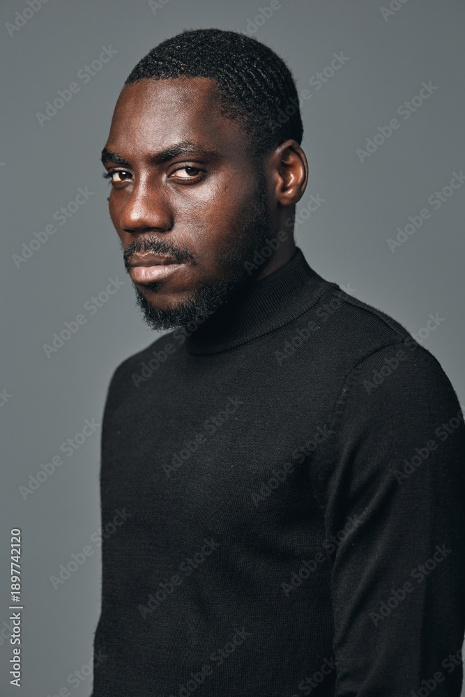 Fototapeta premium African man portrait in creative conceptual visual style wearing black turtleneck sweater with serious expression on gray background. Studio lighting highlights skin texture and facial features.