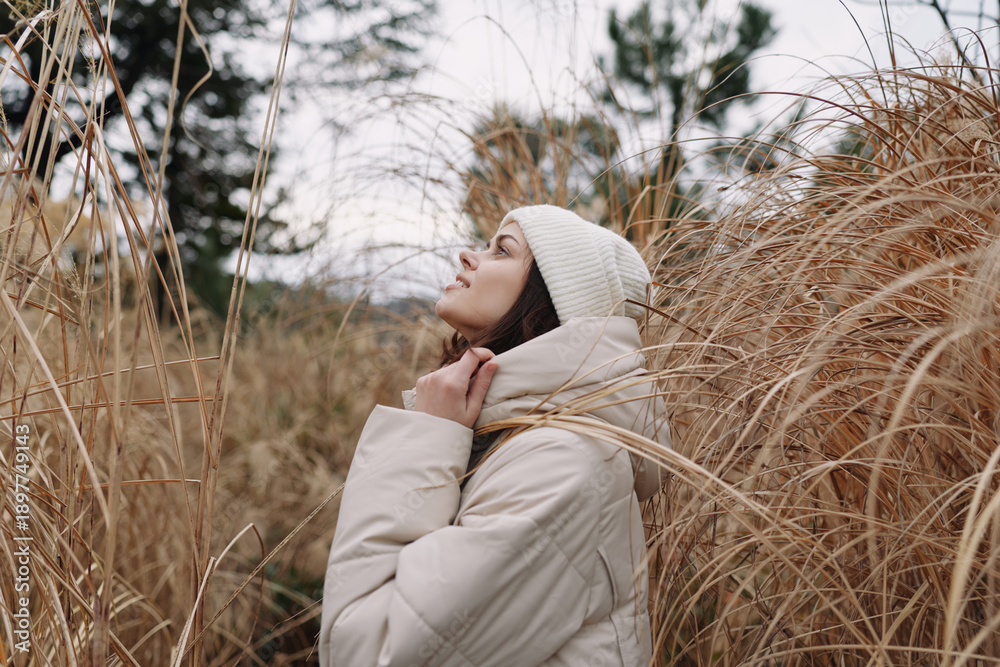 Fototapeta premium Winter portrait of a woman in a beige coat and beanie, standing among tall grass, gazing upward with a serene and reflective mood outdoors.