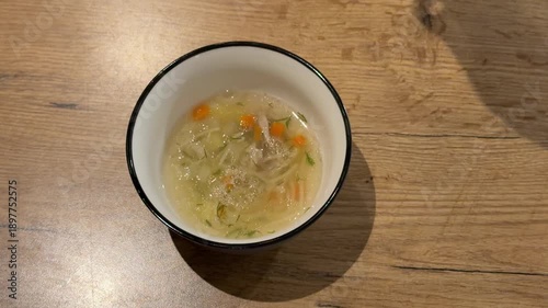 Pouring Vegetable Soup into a Bowl with a Ladle. A ladle pours hot vegetable soup with noodles and carrots into a white bowl on a wooden table, steam rising from the comforting dish.