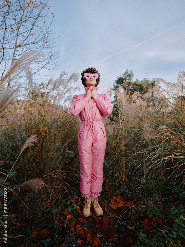 Fototapeta premium Outdoor scene with a person in a pink jumpsuit standing among tall grasses, hands in prayer, wearing a mask, autumnal setting, calm mysterious mood.