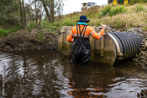 Worker in hi-vis safety gear and waders installs large flood control gate in stream with culvert pipe. Perfect for environmental engineering, water management companies, and infrastructure development