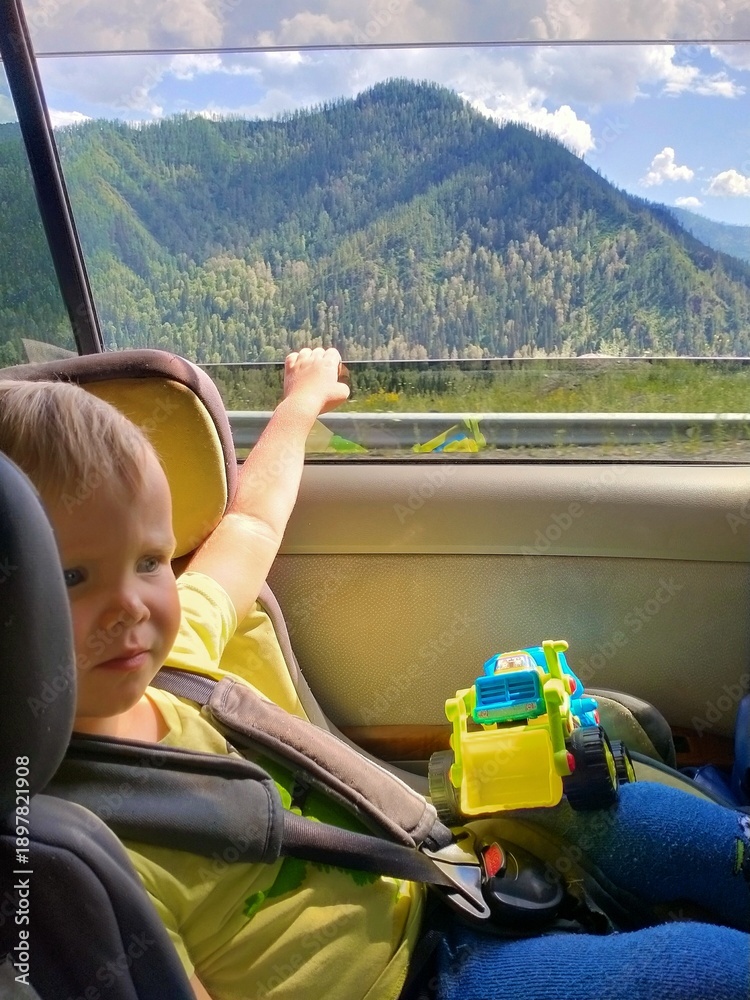 Fototapeta premium A small child sits in a child car seat and looks at the mountains during the trip.