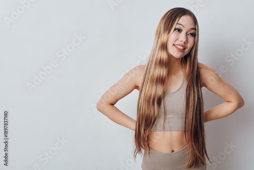 Young woman with long straight hair and casual beige outfit poses smiling with hands on hips against plain light background in studio photography portrait.