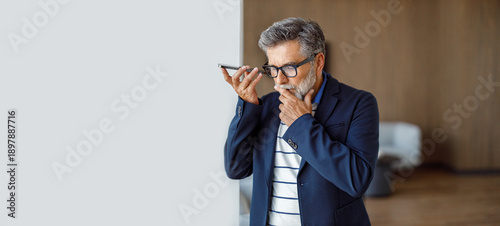 Fotografie Confident businessman in eyeglasses listening to voicemail while standing agains