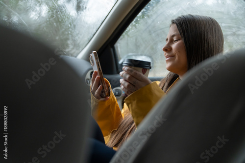 Tablou pe pânză Woman smiling using phone and drinking coffee in car on rainy day